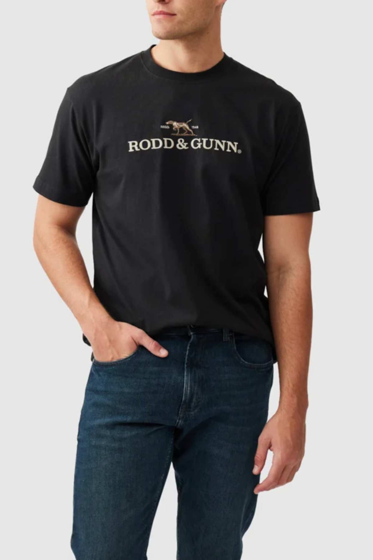 A man wearing the Rodd & Gunn The Peaks Logo Tee, featuring "RODD & GUNN" and an image of a dog printed on the front in black. He complements it with blue jeans and has one hand in his pocket, set against a plain gray background.