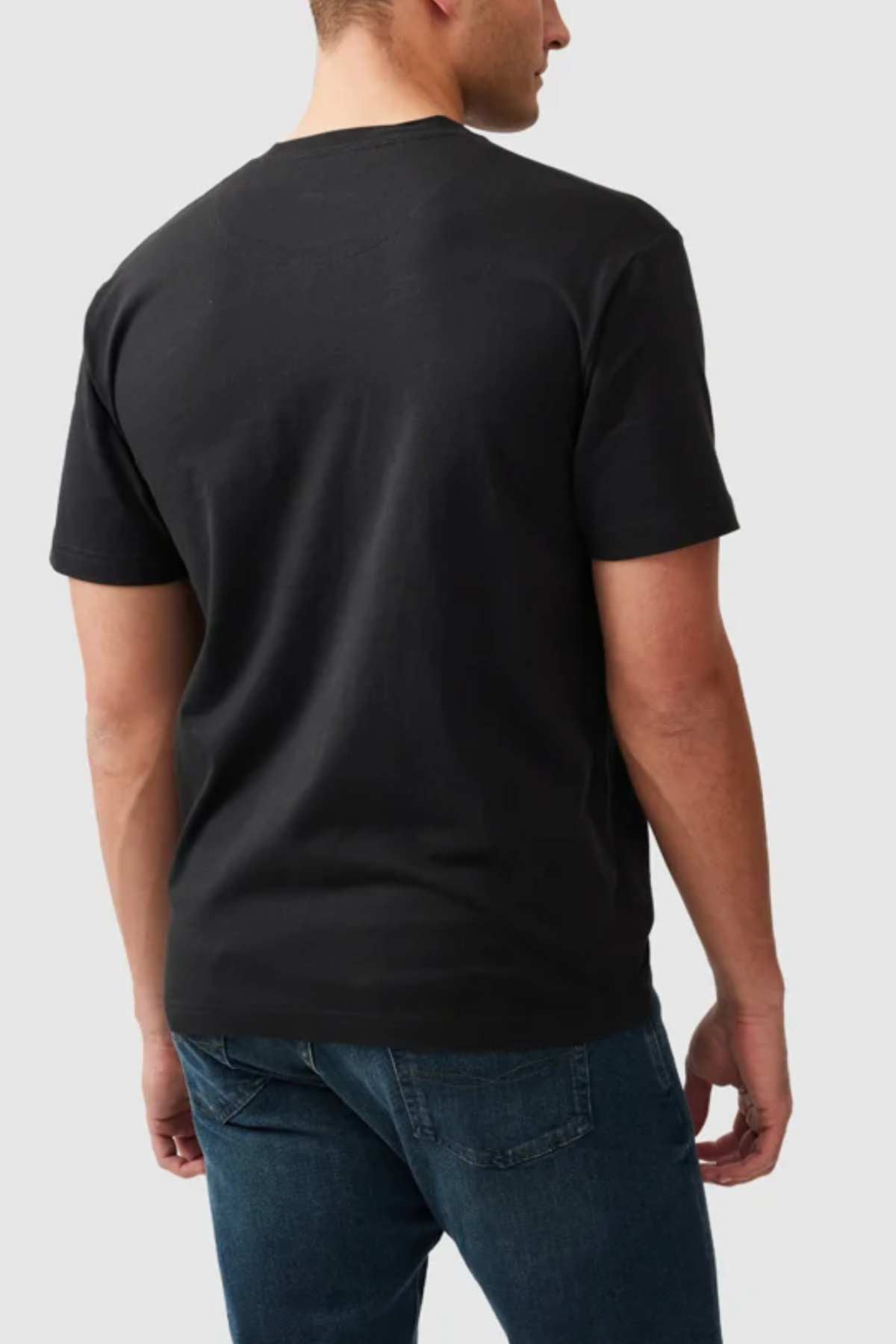 A person with short hair is seen from the back, wearing a Rodd & Gunn The Peaks Logo Tee and blue jeans. The background is plain and light-colored.