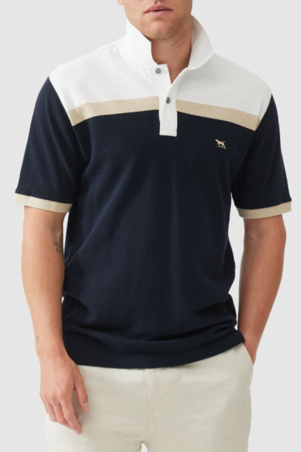 A man wears the Rodd & Gunn Whitby Polo with bold color block design, white and beige accents, and a small embroidered chest logo, paired with light pants, standing with one hand in his pocket.