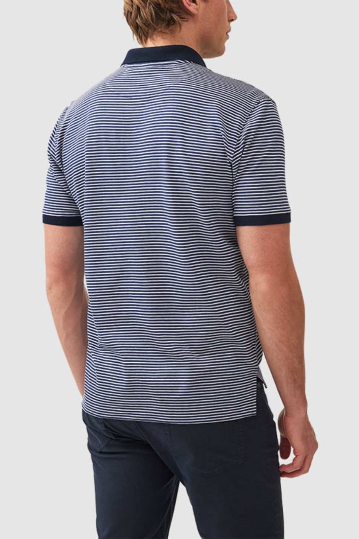 A person with short hair is standing and facing away from the camera. They are wearing a Rodd & Gunn Wye Creek Polo, featuring navy blue and white stripes with dark-colored collar and cuffs, paired with dark pants. The background is plain light gray.