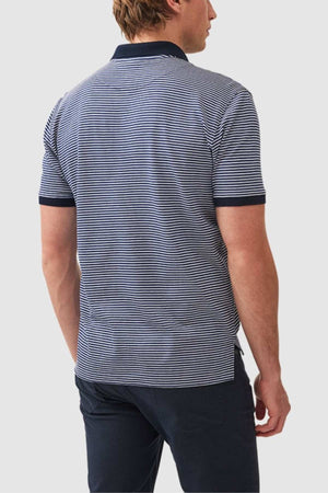 A person with short hair is standing and facing away from the camera. They are wearing a Rodd & Gunn Wye Creek Polo, featuring navy blue and white stripes with dark-colored collar and cuffs, paired with dark pants. The background is plain light gray.