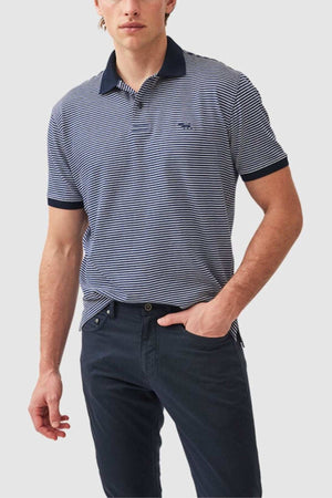 A man wearing the Rodd & Gunn Wye Creek Polo with blue and white stripes, featuring a dark collar and cuffs, paired with dark pants. He has one hand in his pocket and stands against a plain light gray background.