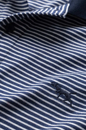 Close-up image of the blue and white striped fabric of the Rodd & Gunn Wye Creek Polo, featuring a small embroidered silhouette of an elephant. The fabric appears to be part of a polo shirt, with visible collar and button details.