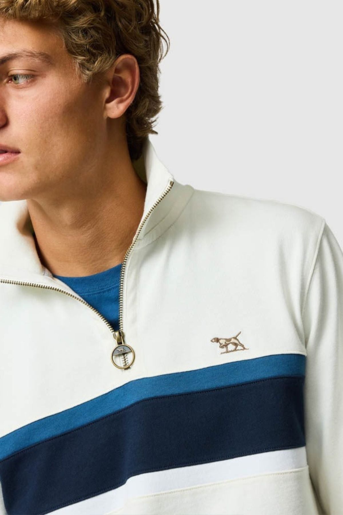 A young person with curly hair wears the Rodd & Gunn Chesterfield Sweat, a cream quarter-zip pullover made from Australian Good Earth Cotton, featuring blue and navy stripes and an embroidered dog logo, layered over a blue shirt.