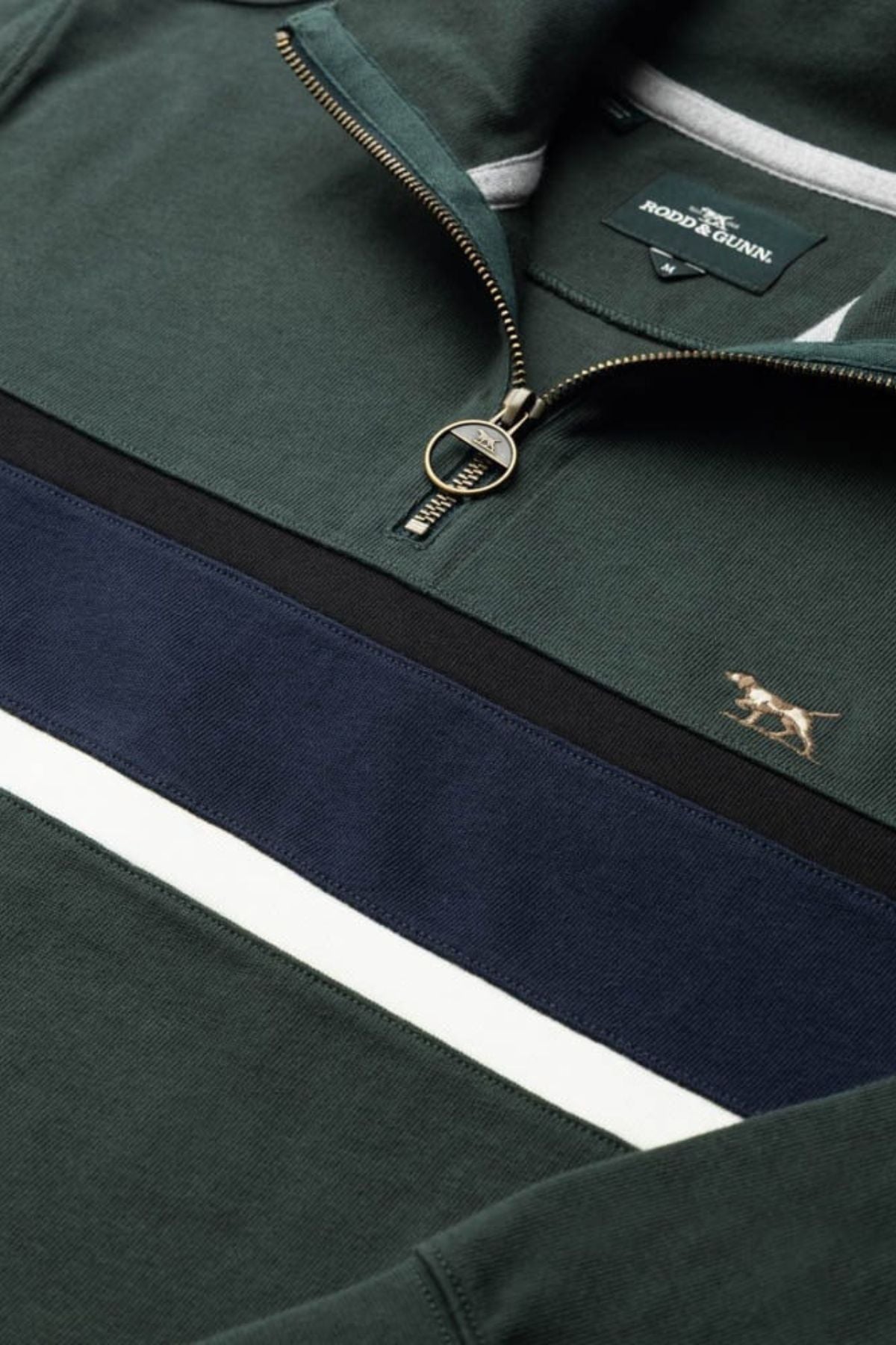 A close-up of the Rodd & Gunn Chesterfield Sweat, a green quarter-zip with a gold dog logo, navy and white stripes, and a metal ring zipper pull. Made with Good Earth Cotton for an eco-conscious style.