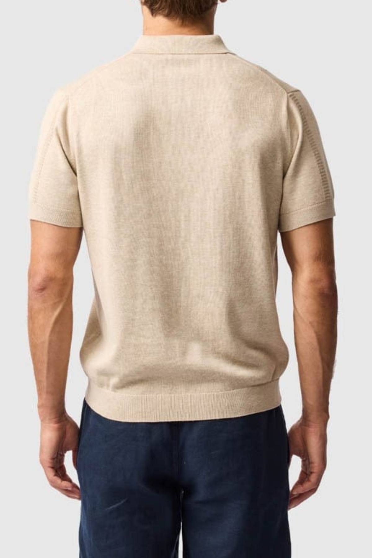 A man wearing the Rodd & Gunn Brooklands Knit, a beige Australian cotton short-sleeve shirt, and dark blue pants is shown from the back against a plain light gray background.