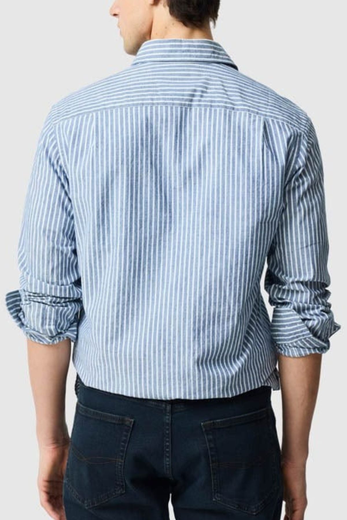 A man is seen from behind wearing the Rodd & Gunn Fernside Sports Fit Shirt, featuring blue and white stripes, rolled-up sleeves, and dark jeans. Made from soft European cotton for a relaxed yet polished appearance.