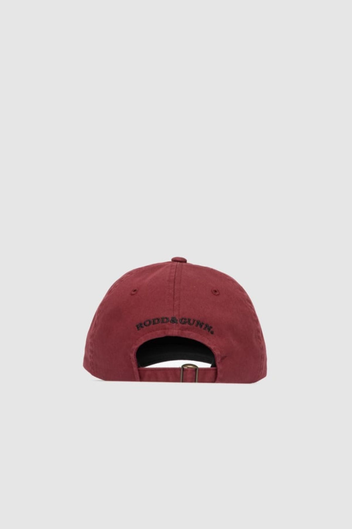The Rodd & Gunn Gunn Cap in maroon cotton twill is shown from the back, featuring a metal buckle strap and "RODD & GUNN" embroidered above the opening. A light grey background highlights its classic style.