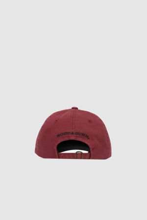 The Rodd & Gunn Gunn Cap in maroon cotton twill is shown from the back, featuring a metal buckle strap and "RODD & GUNN" embroidered above the opening. A light grey background highlights its classic style.