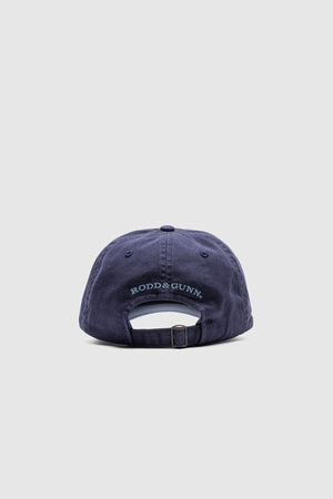 The Rodd & Gunn Gunn Cap, in navy blue cotton twill, is shown from the back with "RODD & GUNN" embroidered above its adjustable strap and buckle. The background is a plain light gray.
