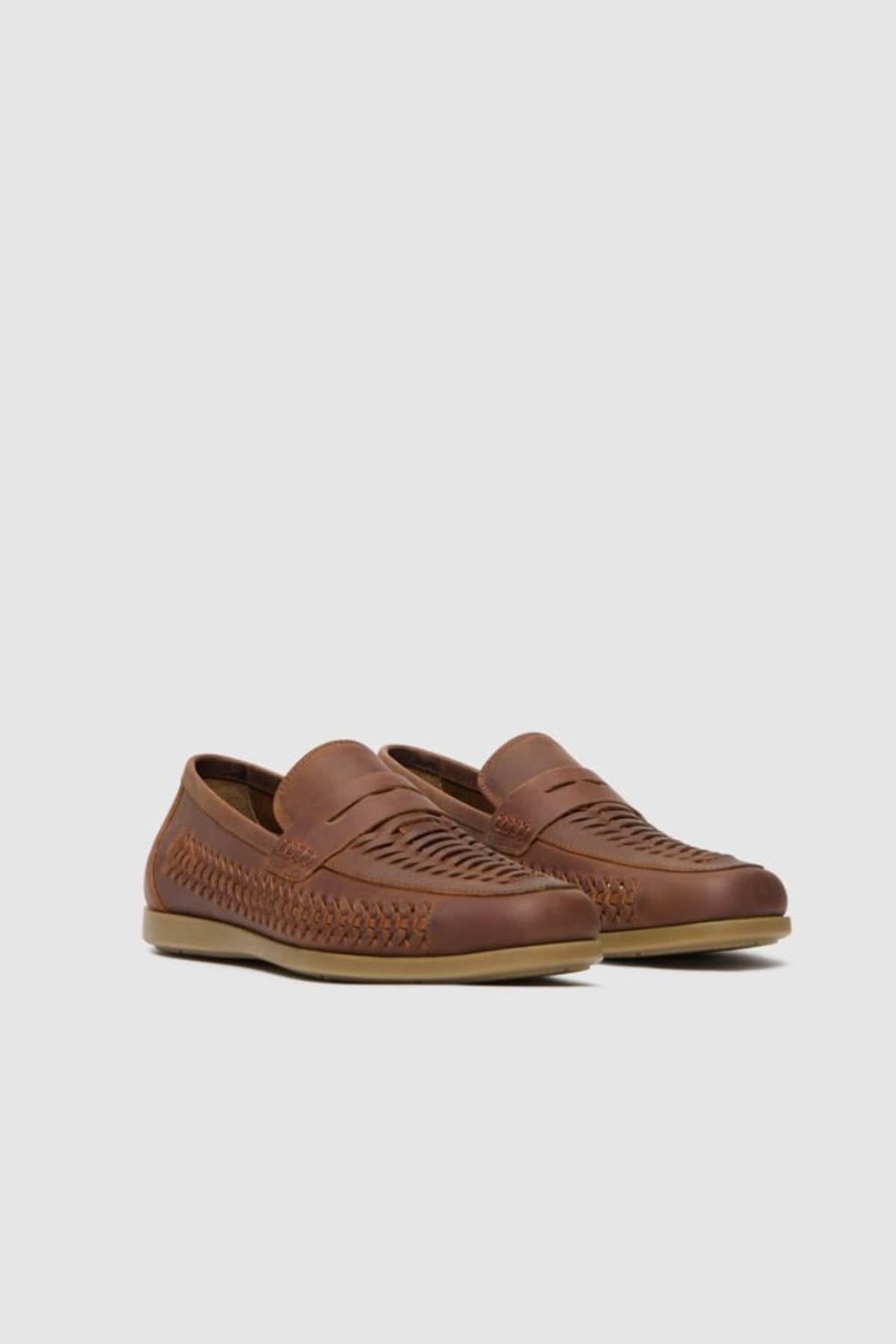 The Rodd & Gunn Gisbourne Huarache Slip On is a brown leather loafer with woven side detailing and a light brown rubber sole for breathable summer comfort, shown against a plain light gray background.