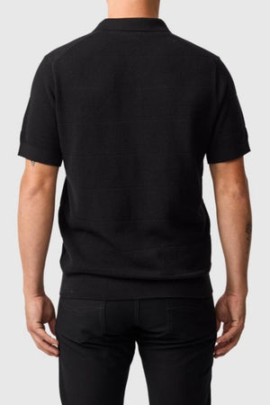 A person is shown from the back, wearing the Rodd & Gunn Masons Flat Knit short-sleeved black cotton shirt and black pants, standing against a plain light gray background.