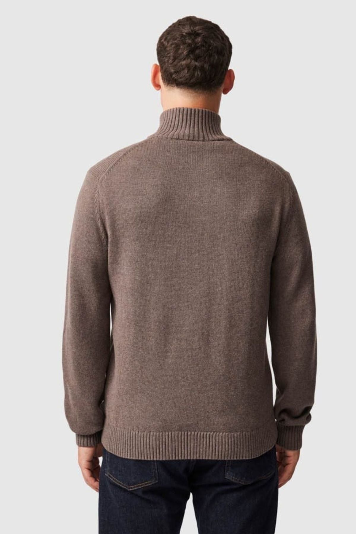 A man is seen from behind, wearing the Rodd & Gunn Merrick Bay Knit—a brown high-neck 100% cotton jersey sweater—with dark blue jeans, set against a plain light gray background.