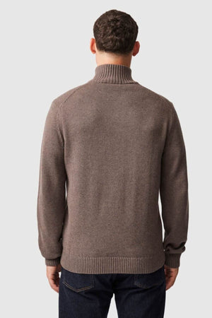 A man is seen from behind, wearing the Rodd & Gunn Merrick Bay Knit—a brown high-neck 100% cotton jersey sweater—with dark blue jeans, set against a plain light gray background.