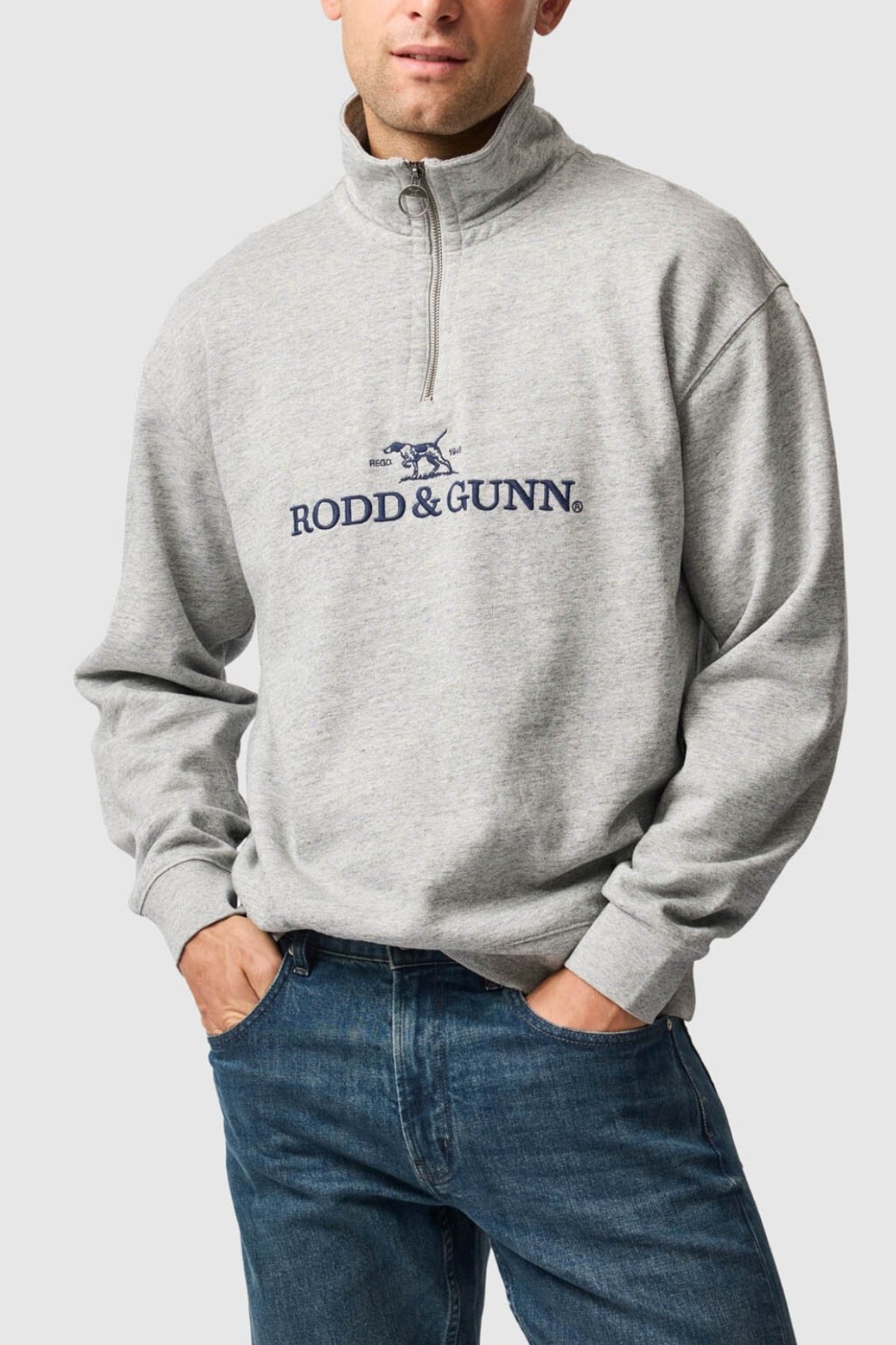 A person wears the Rodd & Gunn Milford Sound Sweat in light gray with "RODD & GUNN" and a dog logo on the front, paired with blue jeans. Its premium YKK metal zip adds refinement as they stand hands in pockets against a plain background.