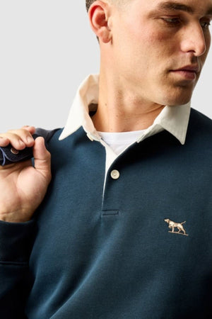 A man wearing a dark blue Rodd & Gunn Otago Sweat with a white collar and embroidered dog logo on the chest looks to the side, holding a strap over his shoulder.