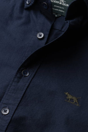 A close-up of the Rodd & Gunn Gunn Oxford Sports Fit Shirt in navy blue, showing a small embroidered dog logo on the chest and part of the inside collar label. Made from 100% cotton for comfort and style.