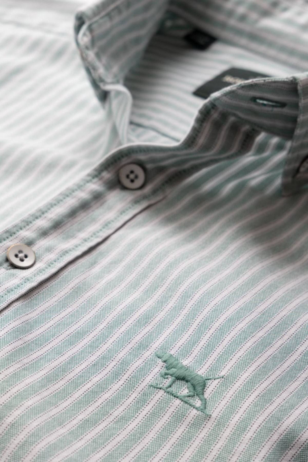 Close-up of the Rodd & Gunn Gunn Oxford Stripe 4 Of Shirt in light blue and white, featuring two buttons, part of the collar, and an embroidered animal logo.