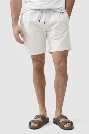 A person is seen from the waist down wearing Rodd & Gunn Resort Shorts with an elastic waist, a light shirt, and brown sandals against a plain light background.