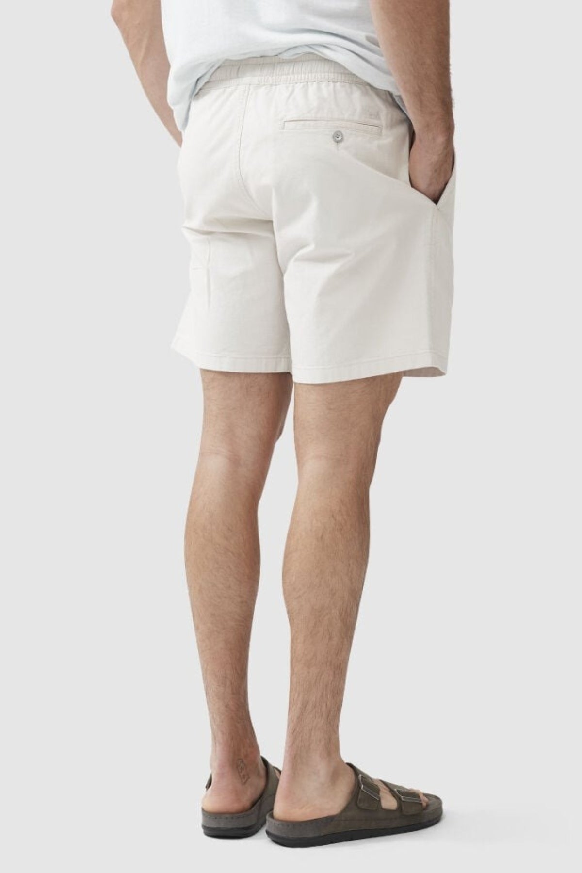 A man stands with his hand in his pocket, wearing Rodd & Gunn Resort Short in a light color, paired with a white shirt and dark sandals, shown from the back against a plain background.