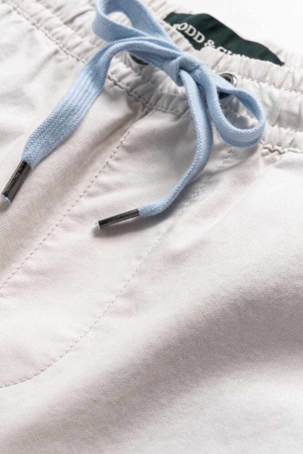 Close-up of Rodd & Gunn Resort Short featuring a visible elastic waistband and light blue drawstring ties with metal tips; part of a green label with white text is seen inside the waistband.