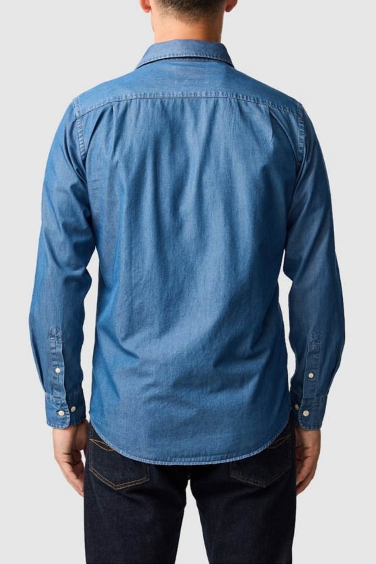 A person stands facing away from the camera in a blue Rodd & Gunn Spring Creek Sports Fit Shirt with cuff buttons and a collar, paired with dark jeans. The background is plain and light-colored.