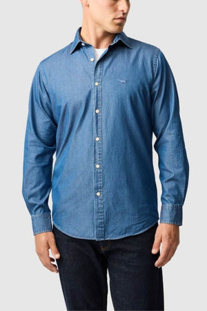 A man wearing a blue Rodd & Gunn Spring Creek Sports Fit Shirt over a white undershirt and dark jeans stands against a plain light gray background.