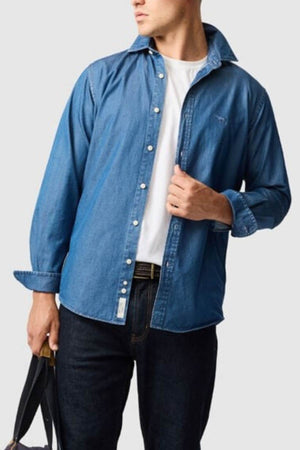 Wearing the Rodd & Gunn Spring Creek Sports Fit Shirt in blue over a white T-shirt and dark jeans, a person stands holding a bag and adjusts their collar against a plain light gray background.