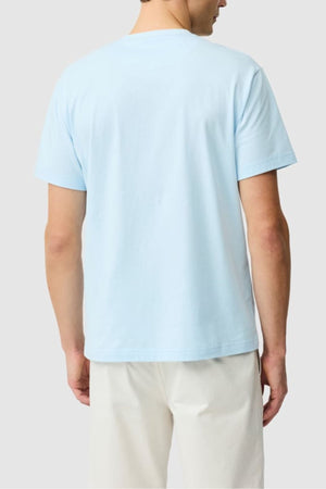 A person faces away from the camera, wearing the Rodd & Gunn The Gunn Tee 2.0 in light blue (100% cotton) and off-white pants, set against a plain light gray background.