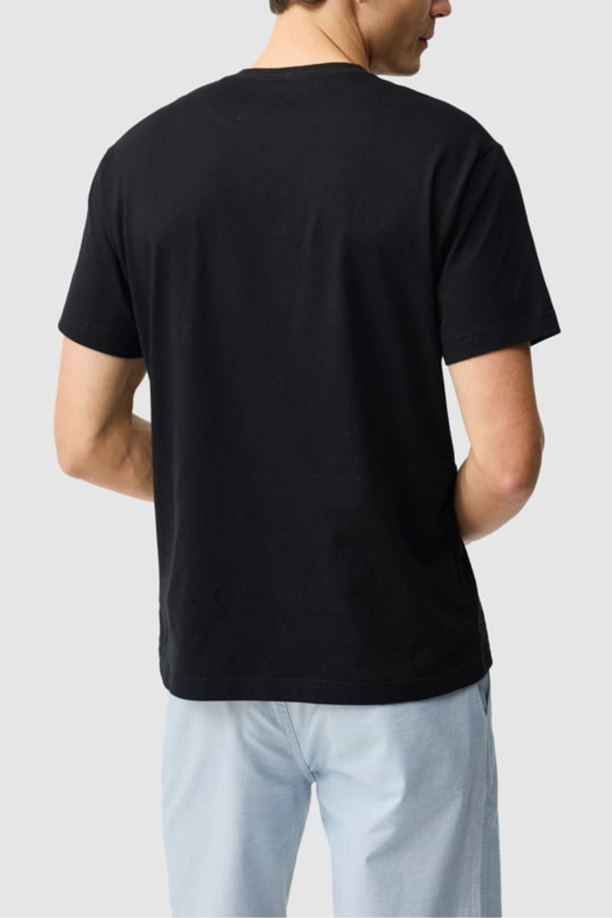 A person is shown from behind wearing the Rodd & Gunn The Gunn Tee 2.0, a plain black 100% cotton sports fit t-shirt, paired with light blue jeans, standing against a light gray background.