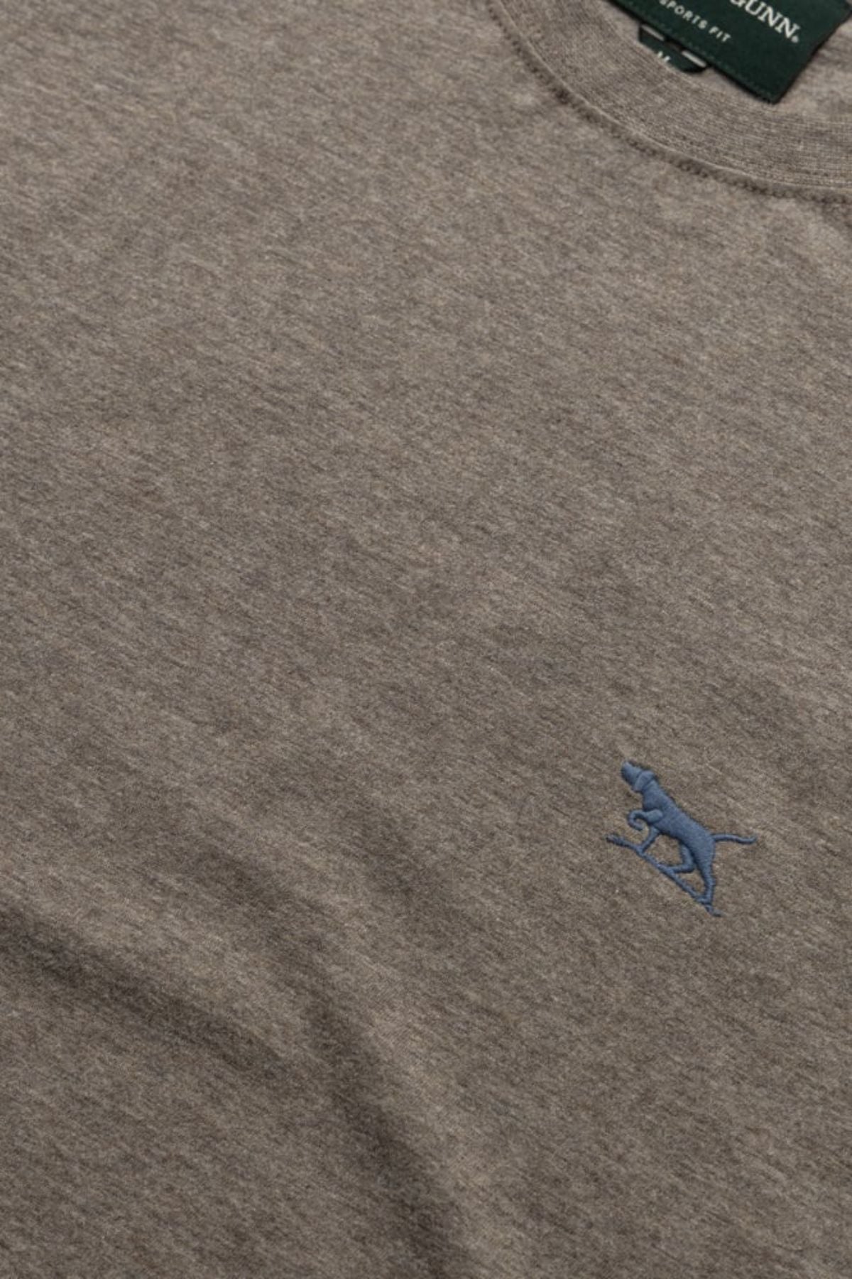A close-up of the heather gray Rodd & Gunn The Gunn Tee 2.0, featuring a small blue embroidered dog logo on the chest, breathable jersey fabric texture, and a glimpse of the green neck tag.