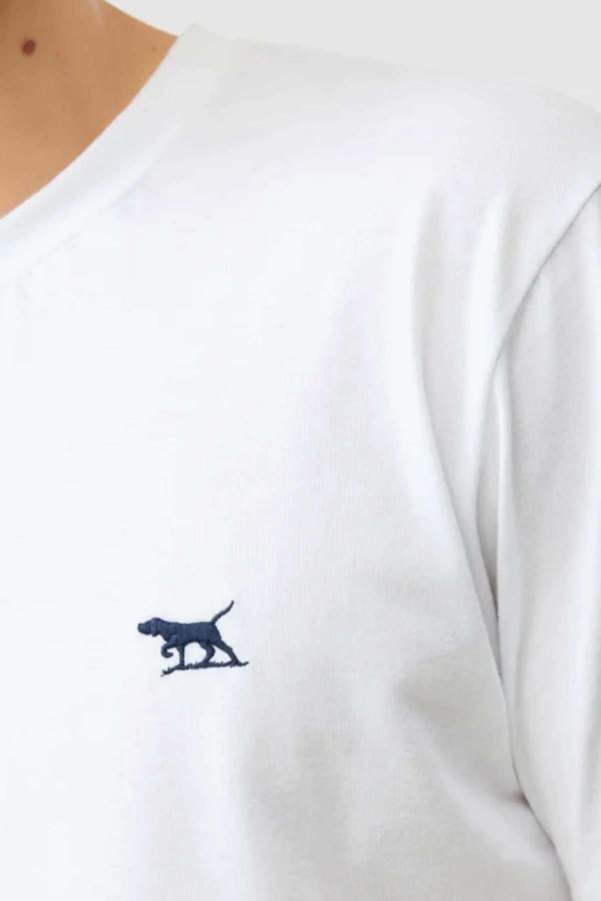 A close-up of a person wearing the white Rodd & Gunn The Gunn Tee 2.0, made from breathable jersey fabric, with a small dark blue embroidered dog logo on the chest. Only part of the neck, shoulder, and upper torso are visible.