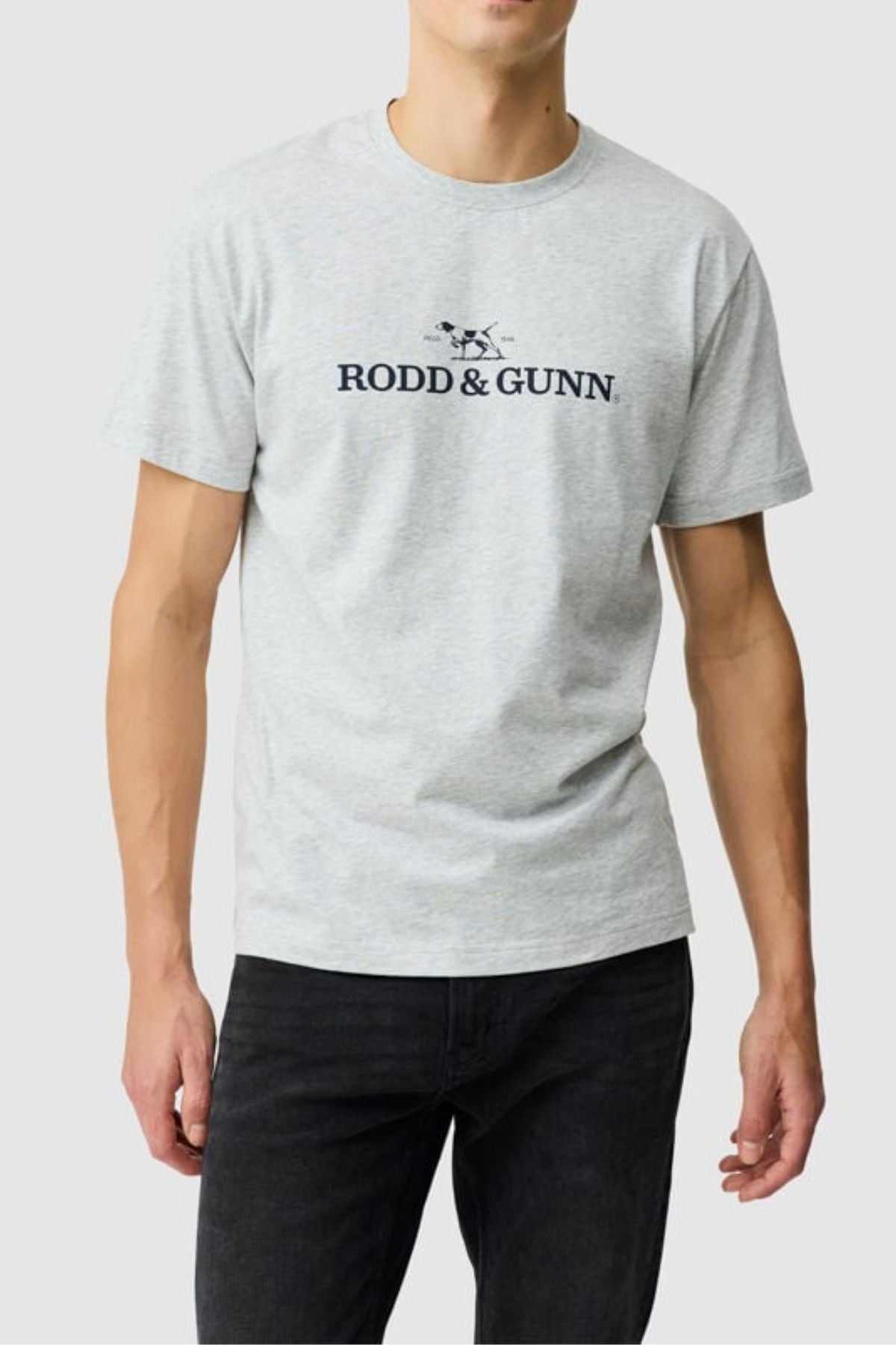 A person wears the Rodd & Gunn The Logo Tee 2.0βa light grey menβs cotton T-shirt with a dog graphic and logoβpaired with black jeans, set against a plain, light background.