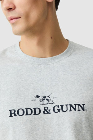 A person wears the light grey Rodd & Gunn The Logo Tee 2.0, a men's cotton t-shirt in a classic sports fit, featuring "RODD & GUNN" and an illustrated dog on the chest, with focus on the chest and a partially visible face.