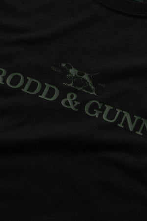A close-up of the Rodd & Gunn The Logo Tee 2.0 in dark cotton, featuring "RODD & GUNN" text, a hunting dog logo, and "REGO 1946"—crafted from 100% cotton for a premium men's sports tee feel.