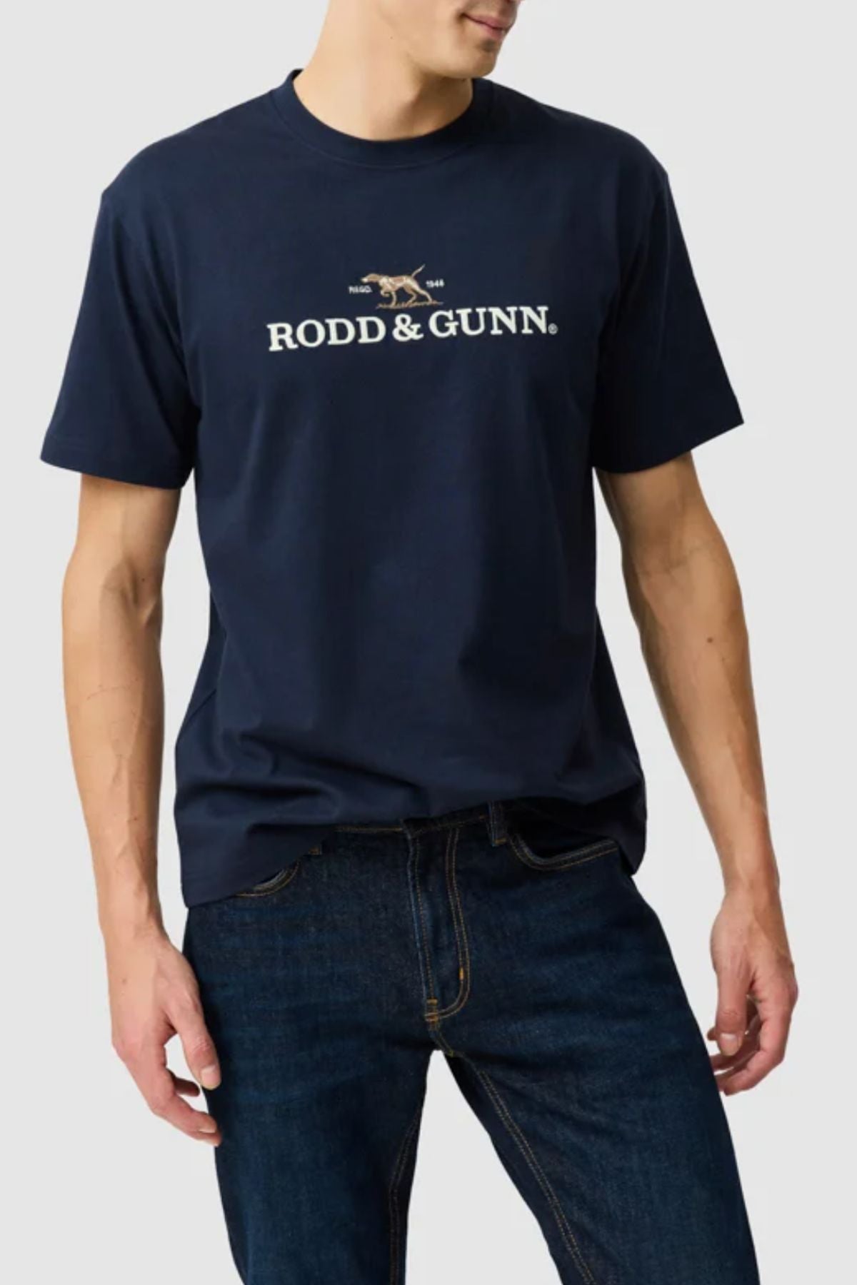 A man wears the Rodd & Gunn The Peaks Logo Tee, featuring FibreTrace technology and a crossed rifles graphic, with dark blue jeans against a light gray background.