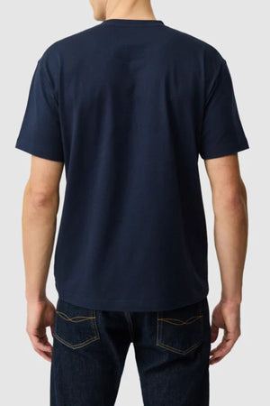 A person in a plain dark blue Rodd & Gunn The Peaks Logo Tee and dark jeans stands with their back to the camera against a light gray background.