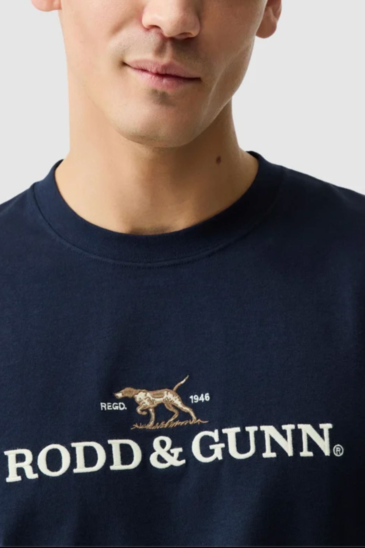 A person wearing a navy blue Rodd & Gunn The Peaks Logo Tee, featuring a hunting dog graphic, "REGD. 1946," and "RODD & GUNN" on the chest, crafted from Australian Good Earth Cotton for comfort and sustainability.