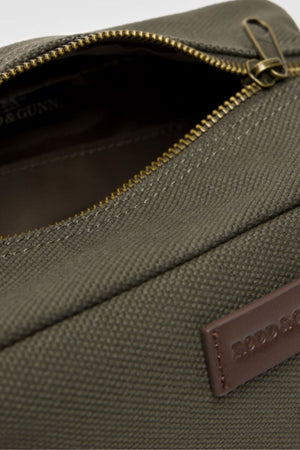 A close-up of the Rodd & Gunn Ponsonby Wash Bag, crafted from olive green cotton canvas with durable textured fabric, a partially open brass zipper, and a brown leather patch label embossed with text.
