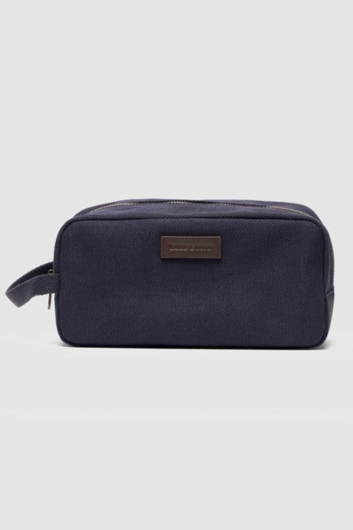 The Rodd & Gunn Ponsonby Wash Bag is a rectangular navy blue cotton canvas bag with a zipper closure, side handle, and a brown patch on the front, shown against a plain light gray background.