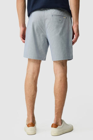 A person facing away stands against a plain light background, wearing the Rodd & Gunn Resort Short in light gray cotton with an elastic waist, paired with a navy shirt and white sneakers with tan accents.
