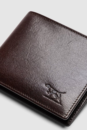A close-up of the Rodd & Gunn Wakefield Bi-Fold Wallet in dark brown Italian leather, featuring an embossed leaping big cat logo on the bottom right, set against a light grey background. This wallet blends classic style with modern craftsmanship.