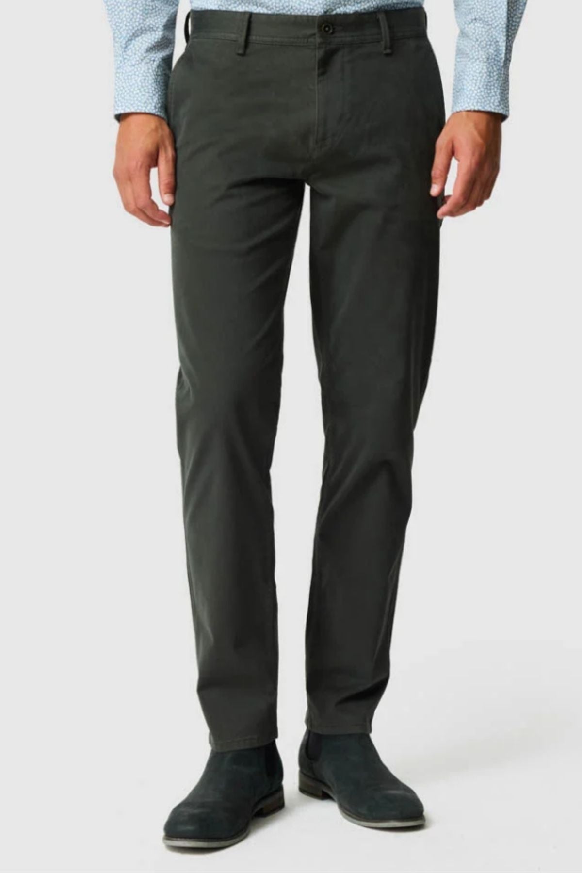 A person wearing Rodd & Gunn Thomas Road 2.0 Chino Pant in dark gray, a light blue patterned dress shirt, and black boots stands against a plain light background. The person's face is not visible.