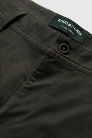 Close-up of Rodd & Gunn Thomas Road 2.0 Chino Pant in dark olive green, featuring a button and pocket detail. The label above the waistband reads "Rodd & Gunn Italian Fabric," highlighting its premium Italian-woven material.