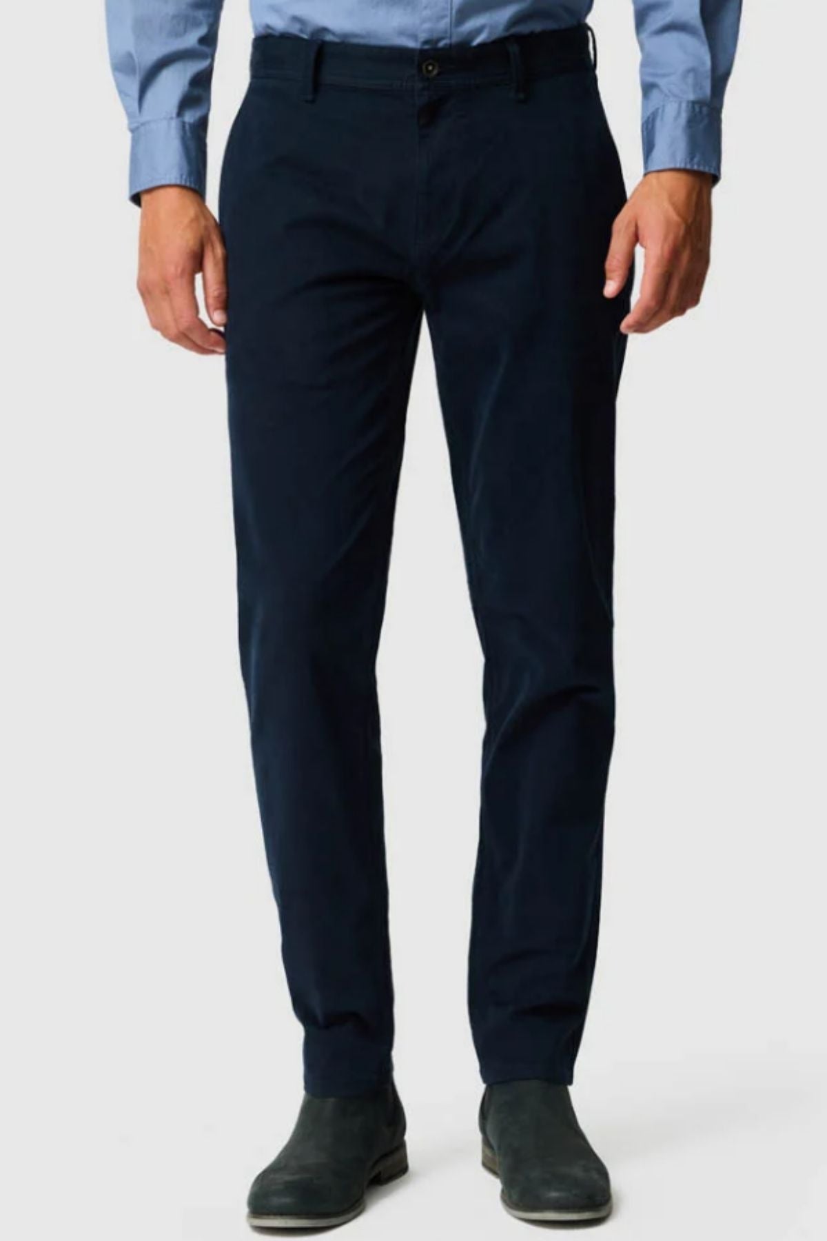 A person wearing a light blue button-up shirt, Rodd & Gunn Thomas Road 2.0 Chino Pant in dark navy, and dark dress shoes stands against a plain light background. Their face is not visible.