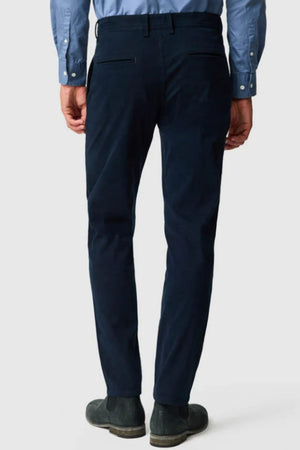 A man is seen from the back, standing against a plain light background, wearing a light blue dress shirt, dark shoes, and Rodd & Gunn Thomas Road 2.0 Chino Pant in dark navy custom fit.