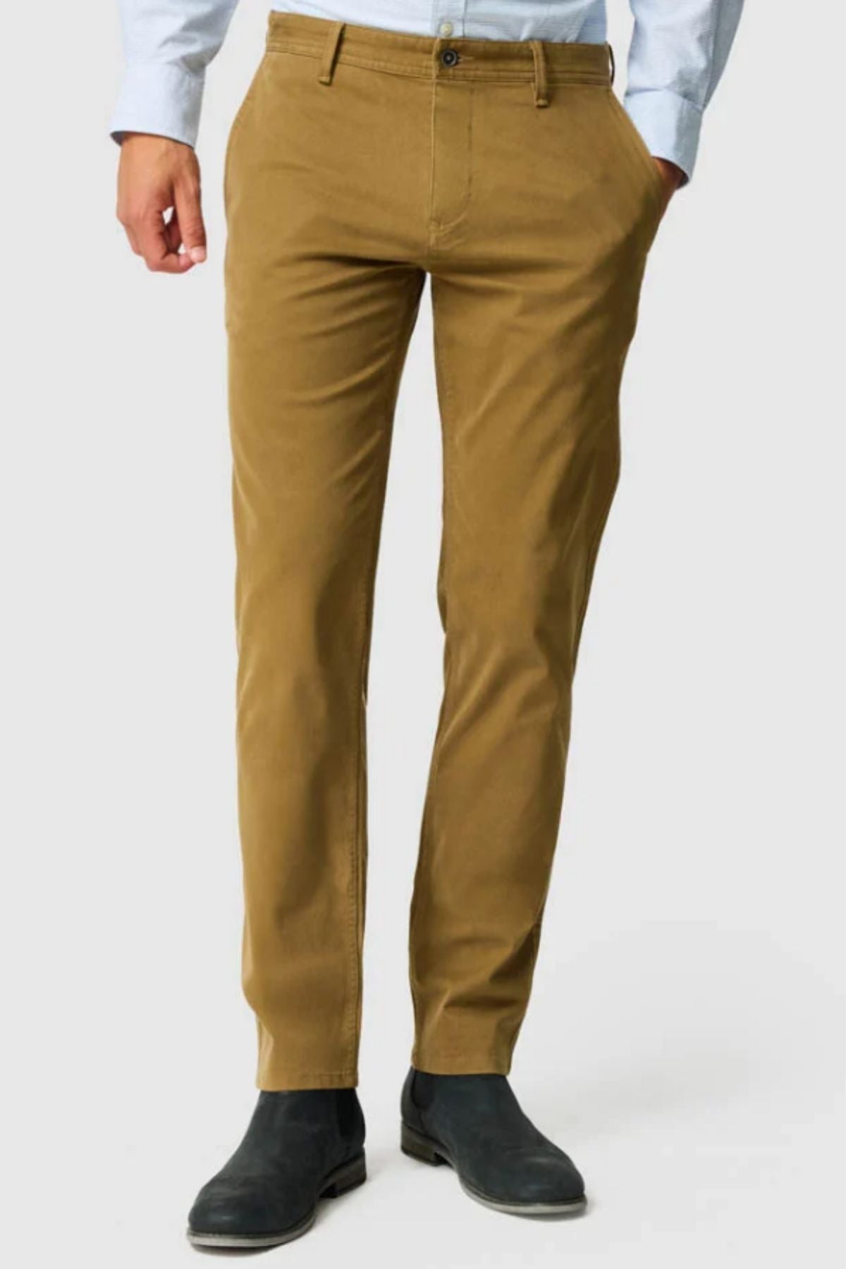 Wearing Rodd & Gunn Thomas Road 2.0 Chino Pant in tan, a light blue shirt, and black ankle boots, a person stands against a plain light background with hands in pockets; the cropped image shows only from the shoulders down.