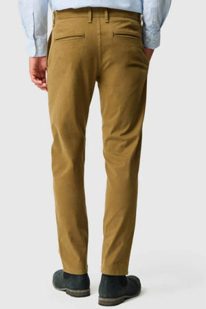 A person wearing Rodd & Gunn Thomas Road 2.0 Chino Pant in light brown, a light blue shirt, and black shoes is standing facing away from the camera against a plain white background.