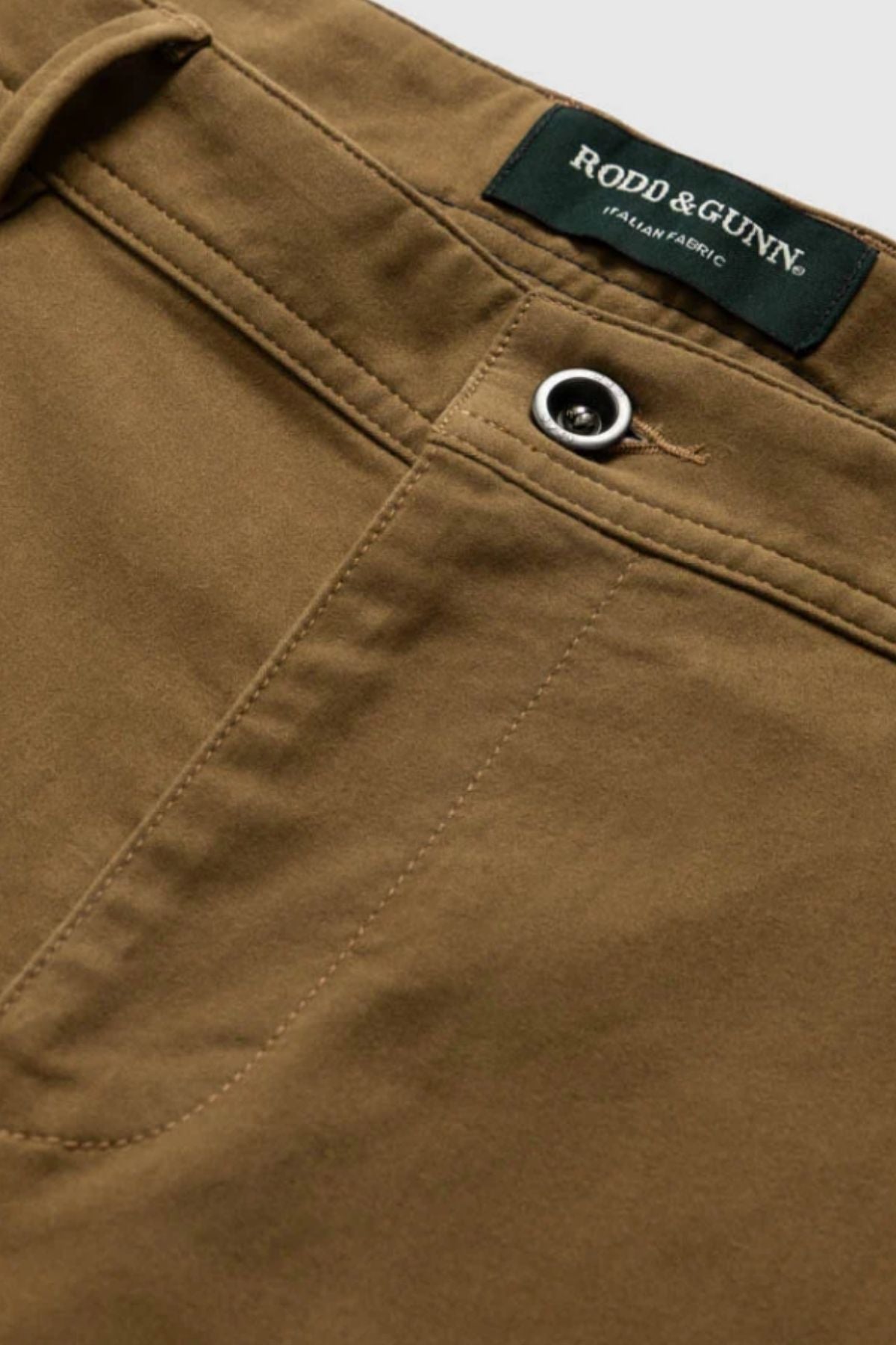 Close-up of the Rodd & Gunn Thomas Road 2.0 Chino Pant in brown, highlighting custom fit, button and stitching details by the waistband, and a green "RODD & GUNN ITALIAN-WOVEN FABRIC" label.