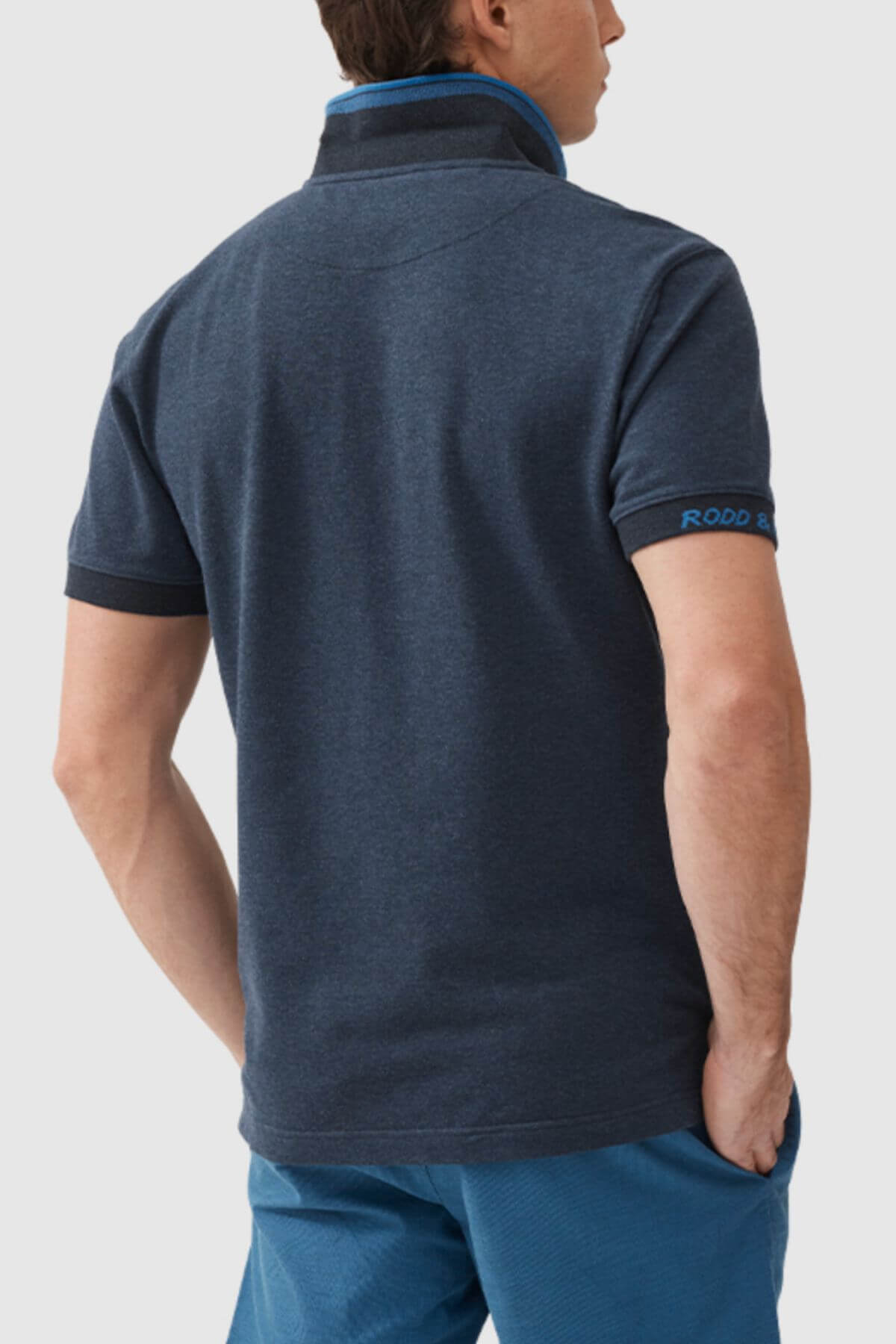 A man, seen from behind, wears a modern slim-fit Rodd & Gunn Masterton Sports Fit Polo in dark blue and blue shorts. His left hand is in his pocket, and the shirt’s right sleeve features blue text. Made from premium USA cotton.